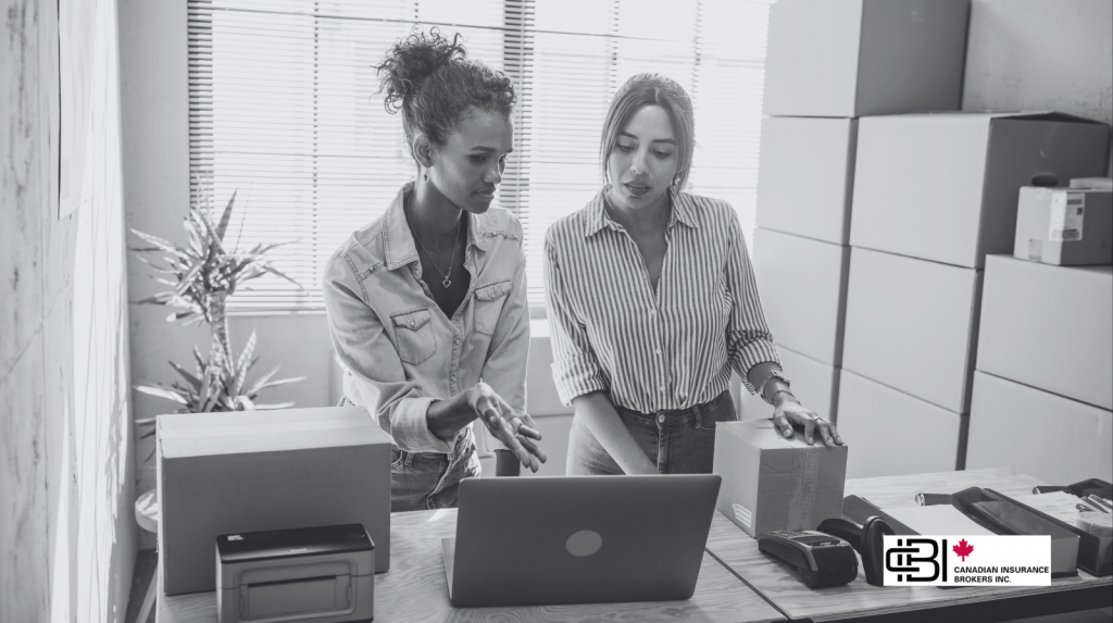 Two small business owners reviewing orders on a laptop while packing boxes in a home workspace and discussing small business insurance in oakville